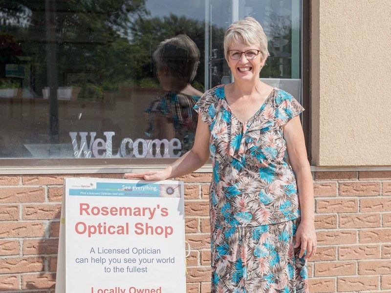 Rosemary standing beside the Rosemary's Optical Shop sign outside the store at 163 Ormond Street, Brockville